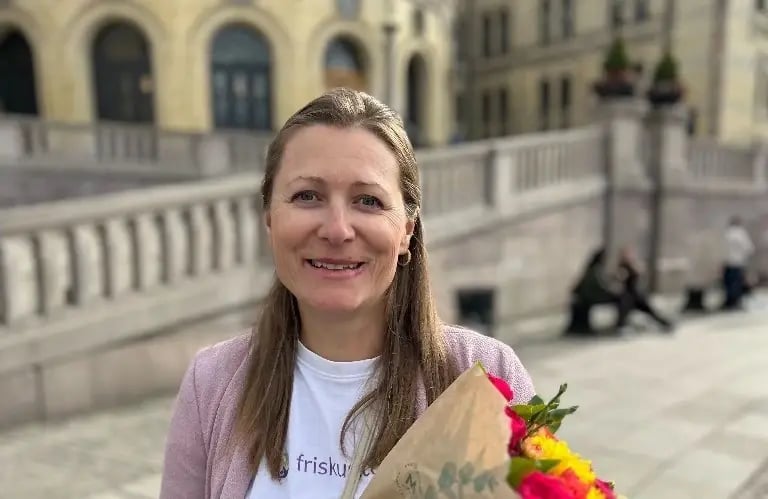 Kvinne står ute med en bukett blomster, smilende mot kameraet, foran en bygning med trapp og balustrade.